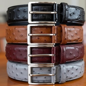 Puede incluir: Una pila de cuatro cinturones de cuero en negro, marr&oacute;n, burdeos y gris. Cada cintur&oacute;n tiene una hebilla plateada y una superficie texturizada con peque&ntilde;as protuberancias espaciadas. Los cinturones est&aacute;n apilados verticalmente, mostrando sus diferentes colores y dise&ntilde;os.