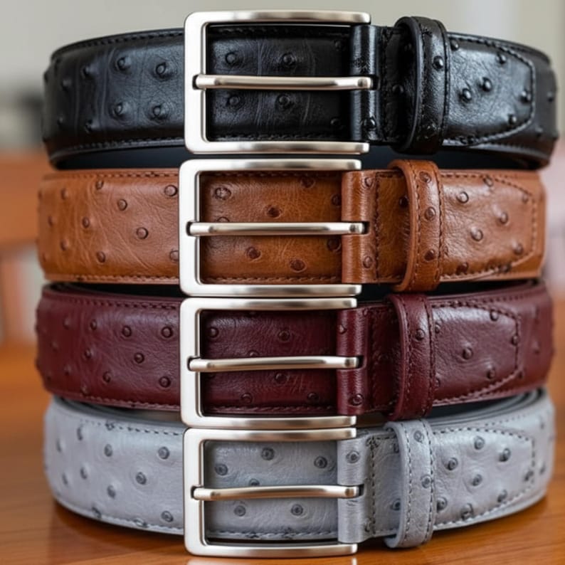 Puede incluir: Una pila de cuatro cinturones de cuero en negro, marr&oacute;n, burdeos y gris. Cada cintur&oacute;n tiene una hebilla plateada y una superficie texturizada con peque&ntilde;as protuberancias espaciadas. Los cinturones est&aacute;n apilados verticalmente, mostrando sus diferentes colores y dise&ntilde;os.