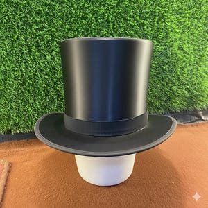 Puede incluir: Un sombrero de copa negro con ala ancha y una banda negra. El sombrero se exhibe sobre un soporte blanco sobre un fondo verde y marrón. Este sombrero clásico es un accesorio formal.