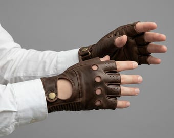May include: A pair of dark brown leather fingerless gloves featuring circular cutouts on the knuckles, perforated detailing on the fingers, and a wrist strap secured with a metallic snap button closure. The gloves are worn over a white shirt sleeve.