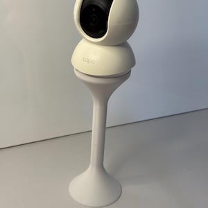 Puede incluir: Cámara de seguridad blanca con lente negra, montada sobre un soporte blanco. La cámara tiene una parte superior redondeada y la marca "tapo" es visible. El soporte tiene una base acampanada y un tallo delgado.