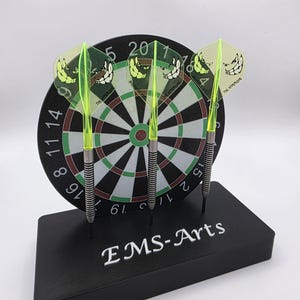 Könnte beinhalten: Ein schwarzes Dartboard mit einem schwarzen Sockel, der drei Darts hält. Die Darts haben neongrüne Schäfte und transparente Flights mit einem Cartoon-Gesicht. Der Sockel trägt den weißen Text "EMS-Arts".