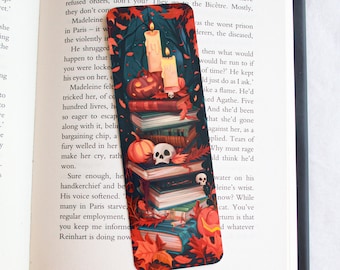 May include: A decorative bookmark featuring a stack of books, pumpkins, skulls, and autumn leaves. Two lit candles and a carved pumpkin sit atop the books. The bookmark is a dark, autumnal design.