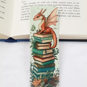 May include: A whimsical bookmark featuring a red dragon perched atop a stack of books. The dragon has detailed scales and wings, with a long tail. The books are various colors, and the bookmark has floral accents.