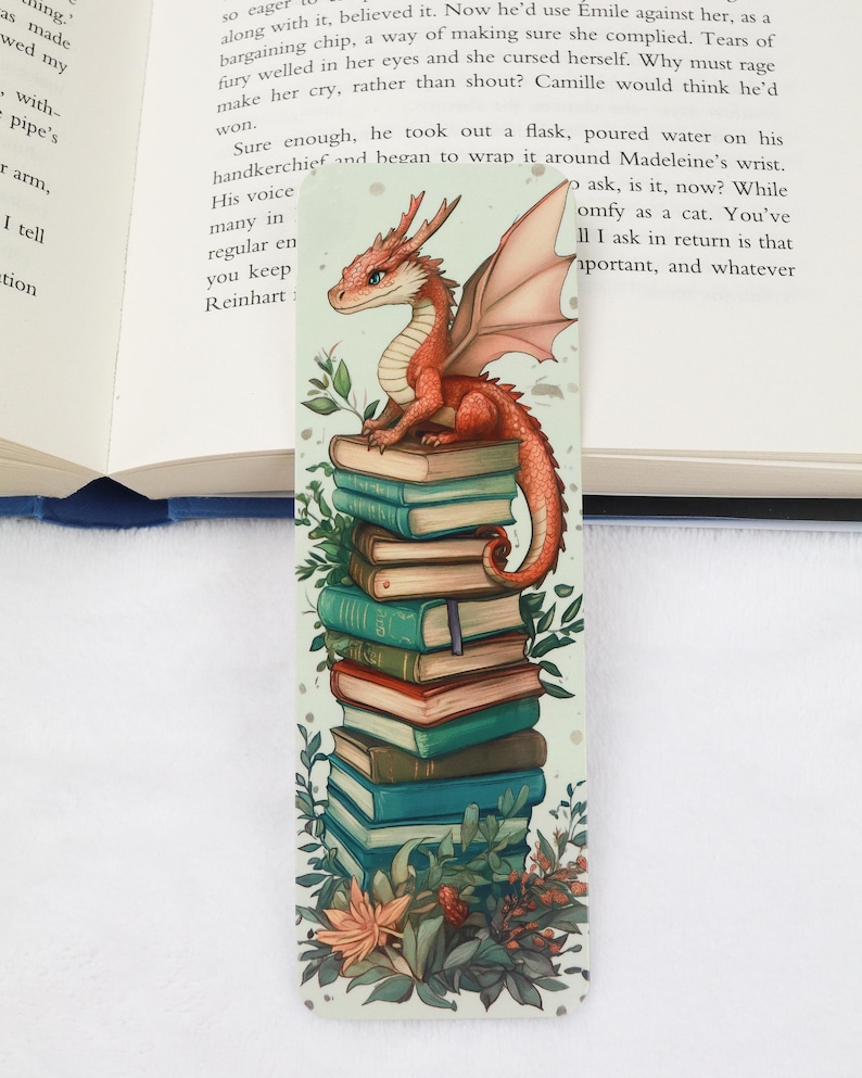 May include: A whimsical bookmark featuring a red dragon perched atop a stack of books. The dragon has detailed scales and wings, with a long tail. The books are various colors, and the bookmark has floral accents.