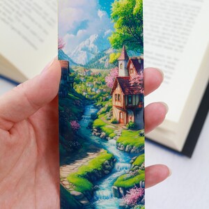 May include: A colorful bookmark featuring a whimsical illustration of a small village nestled in a valley with a winding stream and a charming cottage with a red roof. The bookmark has a blue sky with white clouds and green trees.