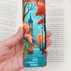 May include: A rectangular bookmark featuring a colourful illustration of a city park path during autumn. The scene includes a red bench, tall street lamps, and trees with orange leaves against a light blue sky and a tall stone building tower.