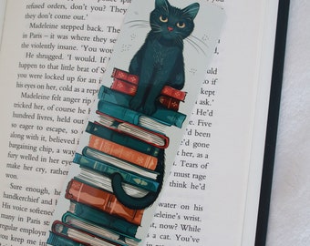 May include: A black cat with green eyes sits on a stack of colorful books. The cat is looking to the right. The text "Well, you" is visible on the page.