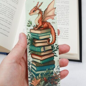 May include: A whimsical bookmark featuring a red dragon perched atop a stack of books. The dragon has detailed wings and a friendly expression. The books are in shades of blue, green, and brown, with botanical accents around the edges. A unique accessory for book lovers.