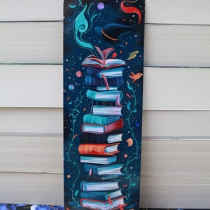 Puede incluir: Un marcapáginas colorido que presenta una pila de libros con un sombrero de bruja y un remolino de magia rojo y verde. El fondo es azul oscuro con estrellas y otros pequeños detalles.