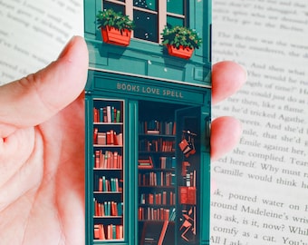 May include: A teal green storefront with a sign that reads "BOOKS LOVE SPELL". The storefront has a large window with a view of a starry night sky. There are two potted plants on the windowsill. The door is open and there are bookshelves filled with books inside.