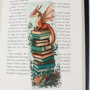 Pode incluir: Marcador de livros com tema de fantasia apresentando um dragão vermelho empoleirado em cima de uma pilha de livros. O dragão tem asas laranja e é cercado por folhagens e flores verdes. O marcador é retangular.