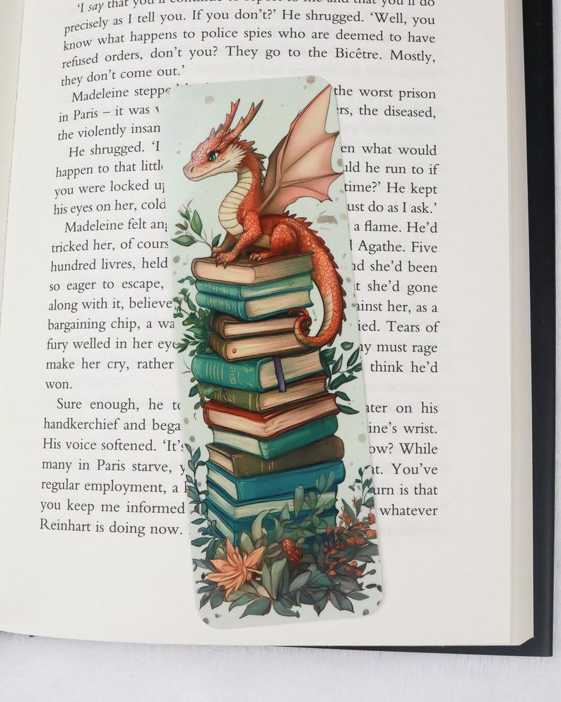 May include: A fantasy-themed bookmark featuring a red dragon perched atop a stack of books. The dragon has orange wings and is surrounded by green foliage and flowers. The bookmark is rectangular and designed for use in books.