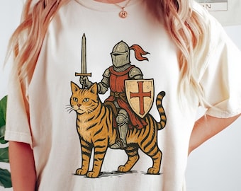 Puede incluir: Camiseta color crema con un caballero de dibujos animados con armadura montado en un gato con rayas de tigre. El caballero sostiene una espada y un escudo con una cruz. El diseño es una versión caprichosa de temas medievales.