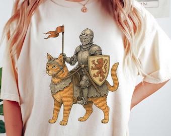 May include: Cream-colored t-shirt featuring a whimsical illustration of a knight in shining armor riding a ginger tabby cat. The knight holds a flag and shield with a lion emblem. The design is detailed with shades of orange, brown, and gray.