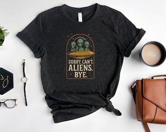 May include: Dark gray t-shirt featuring a graphic of three green aliens in a UFO. The text on the shirt reads "SORRY. CAN'T. ALIENS. BYE." The design is in a rectangular frame with a brown border. The shirt is folded at the bottom.