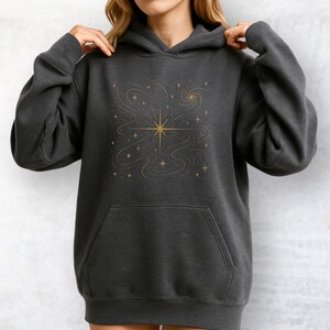 May include: Dark gray hoodie with a gold star and celestial design. The hoodie features a front pocket and a drawstring hood. The gold design includes a central starburst, swirling lines, and smaller stars, creating a cosmic theme.
