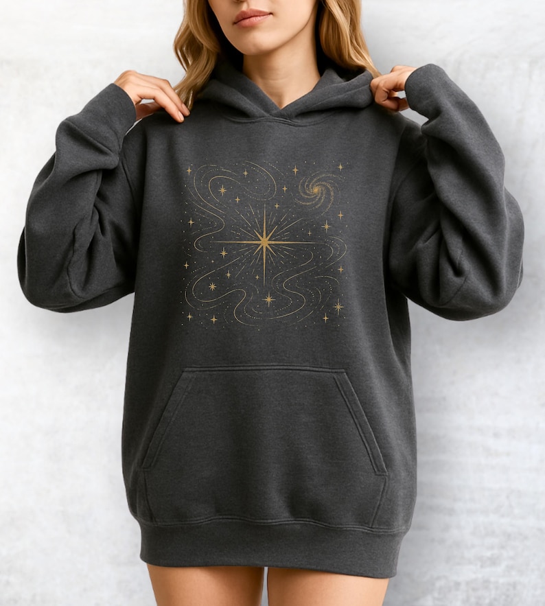 May include: Dark gray hoodie with a gold star and celestial design. The hoodie features a front pocket and a drawstring hood. The gold design includes a central starburst, swirling lines, and smaller stars, creating a cosmic theme.