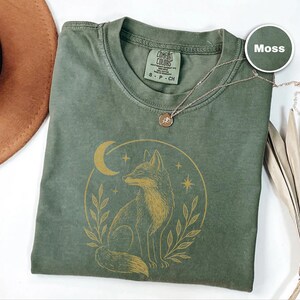 K&ouml;nnte beinhalten: Ein moosgr&uuml;nes T-Shirt mit einem goldenen Fuchsmotiv in einem Mond und Sternen. Das Shirt hat einen Rundhalsausschnitt und kurze &Auml;rmel. Eine zarte Halskette mit rundem Anh&auml;nger liegt auf dem Shirt.