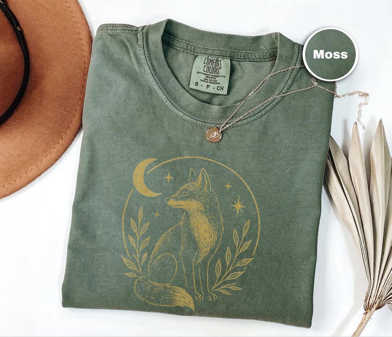 K&ouml;nnte beinhalten: Ein moosgr&uuml;nes T-Shirt mit einem goldenen Fuchsmotiv in einem Mond und Sternen. Das Shirt hat einen Rundhalsausschnitt und kurze &Auml;rmel. Eine zarte Halskette mit rundem Anh&auml;nger liegt auf dem Shirt.