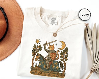 Puede incluir: Camiseta de color marfil con una ilustración caprichosa de un gato caballero a caballo, sosteniendo una espada. El diseño incluye un escudo, una luna, una estrella y árboles. La camiseta tiene una etiqueta Comfort Colors.