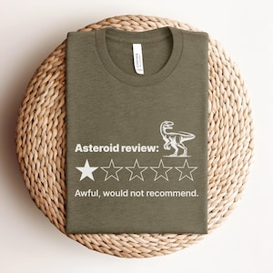 Puede incluir: Camiseta verde oliva con el texto "Asteroid review:" y un dibujo de dinosaurio. La reseña muestra una estrella de cinco y el texto "Awful, would not recommend."