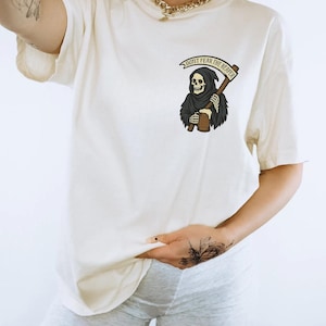 Puede incluir: Camiseta color crema con un gr&aacute;fico de la Parca sosteniendo una guada&ntilde;a y una cerveza. El texto "DON'T FEAR THE REAPER" est&aacute; encima del gr&aacute;fico. La camiseta es de corte holgado.