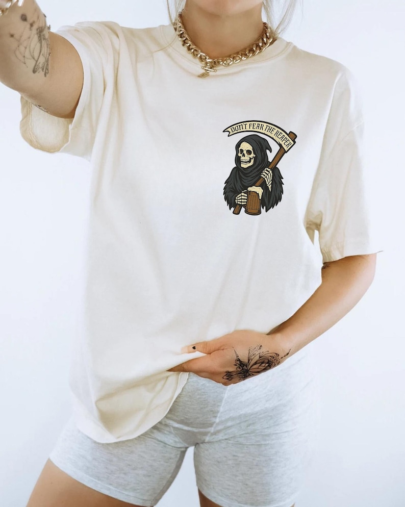 Puede incluir: Camiseta color crema con un gr&aacute;fico de la Parca sosteniendo una guada&ntilde;a y una cerveza. El texto "DON'T FEAR THE REAPER" est&aacute; encima del gr&aacute;fico. La camiseta es de corte holgado.