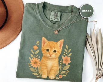 May include: Moss green t-shirt featuring a detailed illustration of an orange kitten surrounded by flowers and leaves. The shirt has a crew neck and short sleeves. A silver necklace with a pendant is on the shirt.