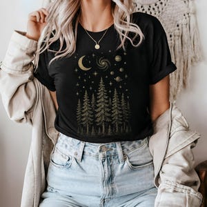 May include: Black t-shirt with a gold celestial design featuring a crescent moon, stars, planets, and a galaxy above a forest of pine trees. The shirt is a crew neck and has short sleeves.