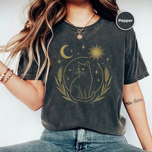 May include: Dark grey t-shirt featuring a gold illustration of a cat, moon, sun, and stars. The cat is centered within a circular design, surrounded by wheat stalks. The shirt has a vintage, worn look.