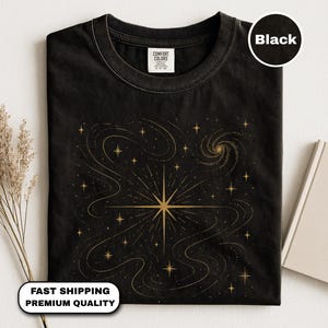 Könnte beinhalten: Schwarzes T-Shirt mit einem goldenen Himmelsdesign, einschließlich eines zentralen Sternenmotivs, wirbelnden Linien und Sternen. Das Shirt hat einen Rundhalsausschnitt und ein "Comfort Colors"-Etikett. Der Text "Black" befindet sich in einem Kreis oben rechts.