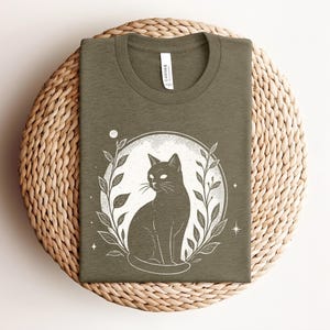 May include: Olive green t-shirt featuring a black cat illustration within a white circle, surrounded by leaves and stars. The shirt is made of soft material and is displayed on a woven surface. The word "CANVAS" is printed on the tag.