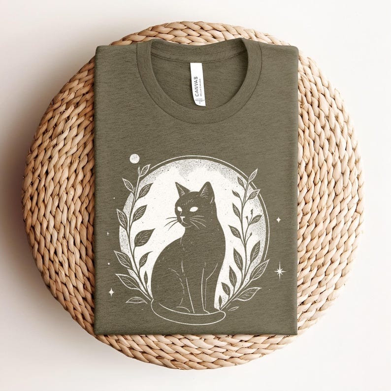 May include: Olive green t-shirt featuring a black cat illustration within a white circle, surrounded by leaves and stars. The shirt is made of soft material and is displayed on a woven surface. The word "CANVAS" is printed on the tag.