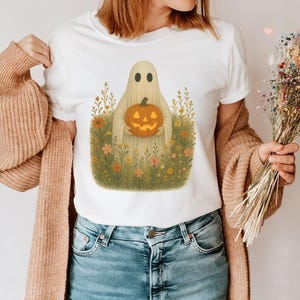 Puede incluir: Camiseta blanca con un fantasma de dibujos animados sosteniendo una calabaza naranja tallada. El fantasma está rodeado de un campo de flores silvestres en tonos verdes, amarillos y naranjas. Ilustración caprichosa de Halloween.