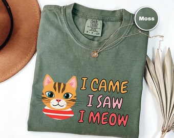 Könnte beinhalten: Moosgrünes T-Shirt mit einem Cartoon-Katzenkopf und dem Text "I CAME I SAW I MEOW" in Orange und Pink. Die Katze hat orangefarbenes Fell, blaue Augen und einen rot-weiß gestreiften Kragen. Eine goldene Halskette liegt auf dem Shirt.