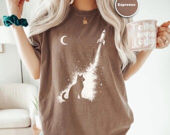 May include: Espresso-colored t-shirt featuring a cat silhouette, a rocket launching into space, and a crescent moon. The graphic is in white. The shirt is a relaxed fit.