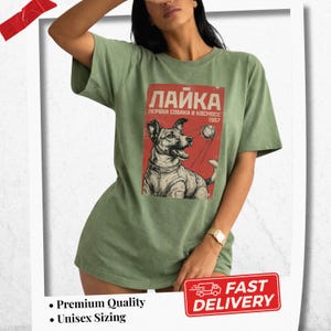 Könnte beinhalten: Salbeigrünes T-Shirt mit einem Retro-Grafikdesign eines Hundes im Raumanzug, dem russischen Text "Laika" und "Erster Hund im Weltraum 1957" auf rotem Hintergrund. Das T-Shirt hat einen Rundhalsausschnitt und kurze Ärmel.