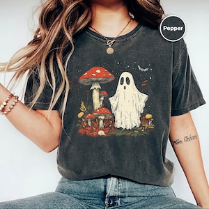Può includere: T-shirt grigio scuro con un'illustrazione di un fantasma e funghi. Il design presenta un fantasma amichevole e diversi funghi con cappelli rossi, ambientati in un fogliame autunnale. Lo stile generale è stravagante e leggermente spettrale.