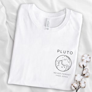 May include: White t-shirt with a black graphic of Pluto, a planet with a line drawing of its surface. The text "Pluto" is above the graphic and "Never Forget 1930-2006" is below.