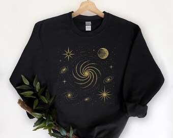 Peut inclure: Sweat-shirt noir avec un motif galaxie doré. Le motif représente une galaxie spirale centrale, des étoiles et des planètes. Le sweat-shirt est suspendu à un cintre en bois sur fond blanc.