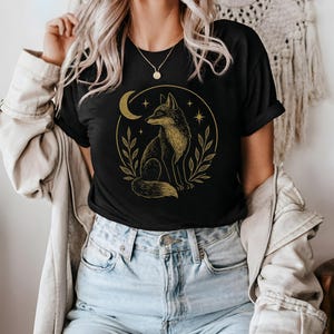 Könnte beinhalten: Schwarzes T-Shirt mit einer goldenen Fuchsgrafik in einem Kreis, einschließlich Mond, Sternen und Blättern. Das T-Shirt besteht aus weichem Material und eignet sich für den Freizeitgebrauch. Das Design ist auf der Vorderseite aufgedruckt.