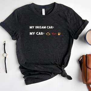 May include: Dark grey t-shirt with the text "MY DREAM CAR:" above "MY CAR:" and car dashboard symbols. The shirt is knotted at the bottom. A watch, glasses, and a brown handbag are visible.