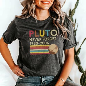 May include: A dark grey t-shirt with a vintage-style graphic that says "Pluto Never Forget 1930-2006" with a rainbow stripe design and a cartoon image of Pluto.