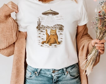 Könnte beinhalten: Weißes T-Shirt mit einem humorvollen Aufdruck einer orangefarbenen Tabby-Katze in einer Wüstenlandschaft mit einem UFO darüber. Die Katze ist in einer komischen Pose dargestellt. Das Design verwendet Braun- und Beigetöne.
