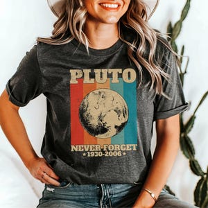Könnte beinhalten: Ein dunkelgraues T-Shirt mit einer Retro-Grafik von Pluto, einem Planeten mit einem Regenbogenhintergrund. Der Text "Pluto Never Forget *1930-2006*" ist auf dem Shirt aufgedruckt.