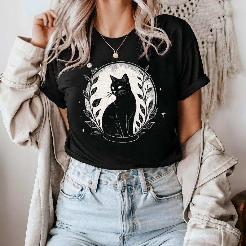 May include: Black t-shirt featuring a black cat illustration within a white circle, surrounded by leaves and stars. The cat is sitting and looking to the side. The design is printed on a black t-shirt.