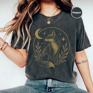 K&ouml;nnte beinhalten: Dunkelgraues T-Shirt mit einer goldenen Illustration eines Fuchses in einem Kreis, zusammen mit einem Mond, Sternen und Bl&auml;ttern. Das Shirt hat einen Vintage-Look. Das Wort "Pepper" befindet sich in einem Kreis oben rechts.