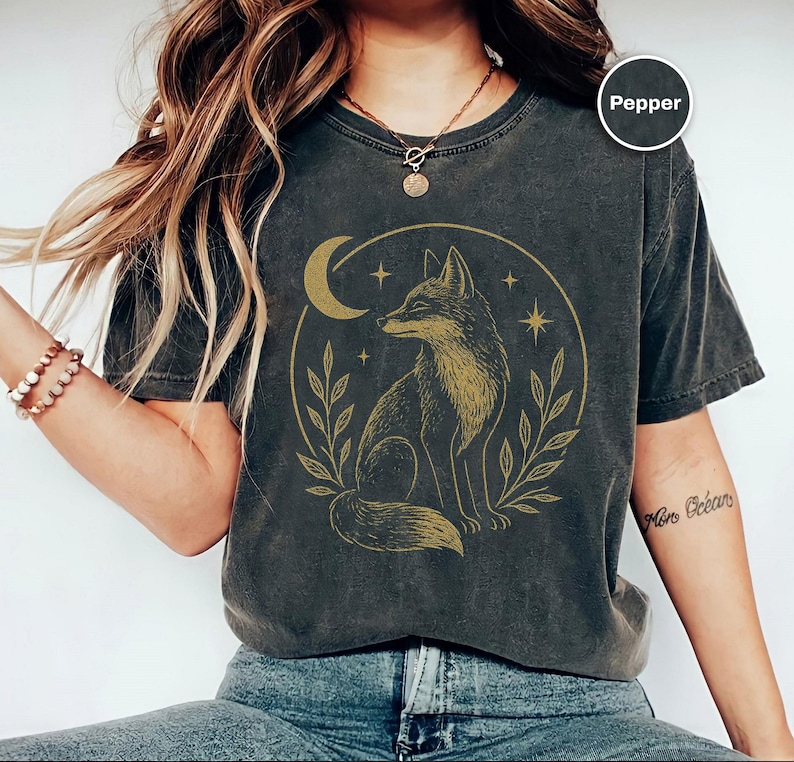 K&ouml;nnte beinhalten: Dunkelgraues T-Shirt mit einer goldenen Illustration eines Fuchses in einem Kreis, zusammen mit einem Mond, Sternen und Bl&auml;ttern. Das Shirt hat einen Vintage-Look. Das Wort "Pepper" befindet sich in einem Kreis oben rechts.