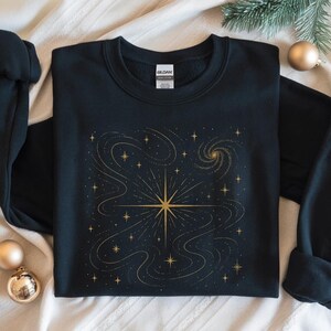 May include: Black sweatshirt featuring a gold celestial design. The graphic includes a central starburst, swirling lines, and numerous smaller stars. The design is centered on the front of the long-sleeved garment, creating a cosmic theme.